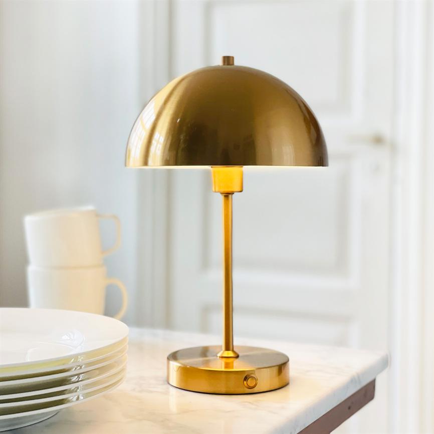 En guldfarvet bordlampe står tændt på et marmorbord; rund skærm over slank stang, hvide kaffekopper og en stak tallerkener i et lyst køkkenmiljø.