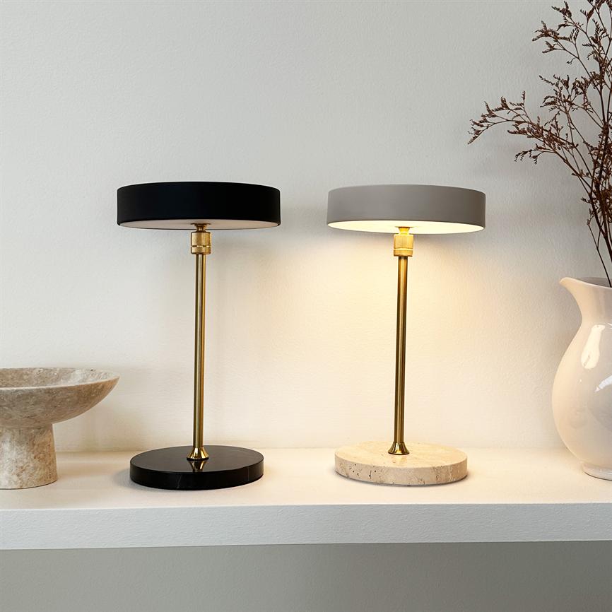 To bordlamper står side om side; den højre lyser svagt, den venstre er slukket; begge med messingstandere på en hvid hylde mod en lys væg, flankeret af skål og vase.