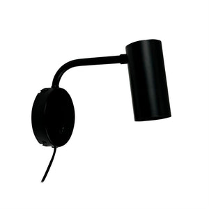 Sort metal væglampe monteret på rund vægplade, arm bøjet udad og cylinderformet lampehoved peger skråt nedad; enkel, moderne design mod hvid baggrund.