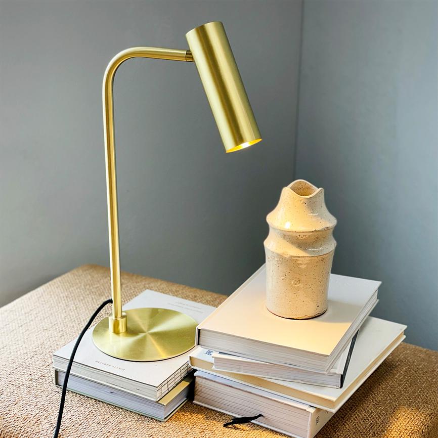 Guldfarvet bordlampe lyser ned over en stak hvide bøger, hvor en beige, skulpturel keramikvase står; motivet står på en fletoverflade mod en grå væg.