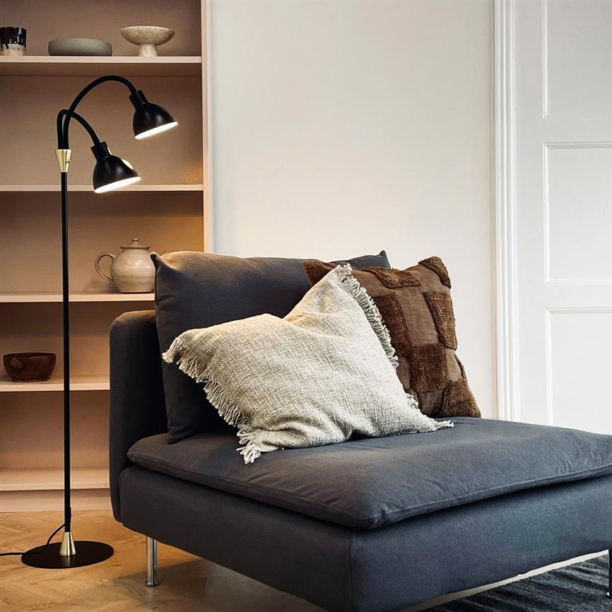 En mørkegrå chaiselong hviler med to dekorative puder, en lysegrå med frynser og en brun ternet; en sort gulvlampe lyser svagt ved hylder med keramik i et lyst, minimalistisk rum.