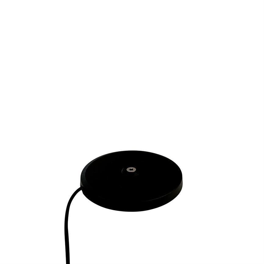 Sort, rund opladningsbase med metalstik i midten ligger fladt, et sort kabel fører væk; genstanden er isoleret mod en ensartet hvid baggrund.