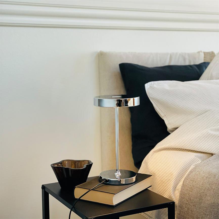 Bordlampe står på en bog på et sort natbord; en mørk skål ved siden af; natbordet står ved en beige seng med lyse, stribede sengetøj og en sort pude.