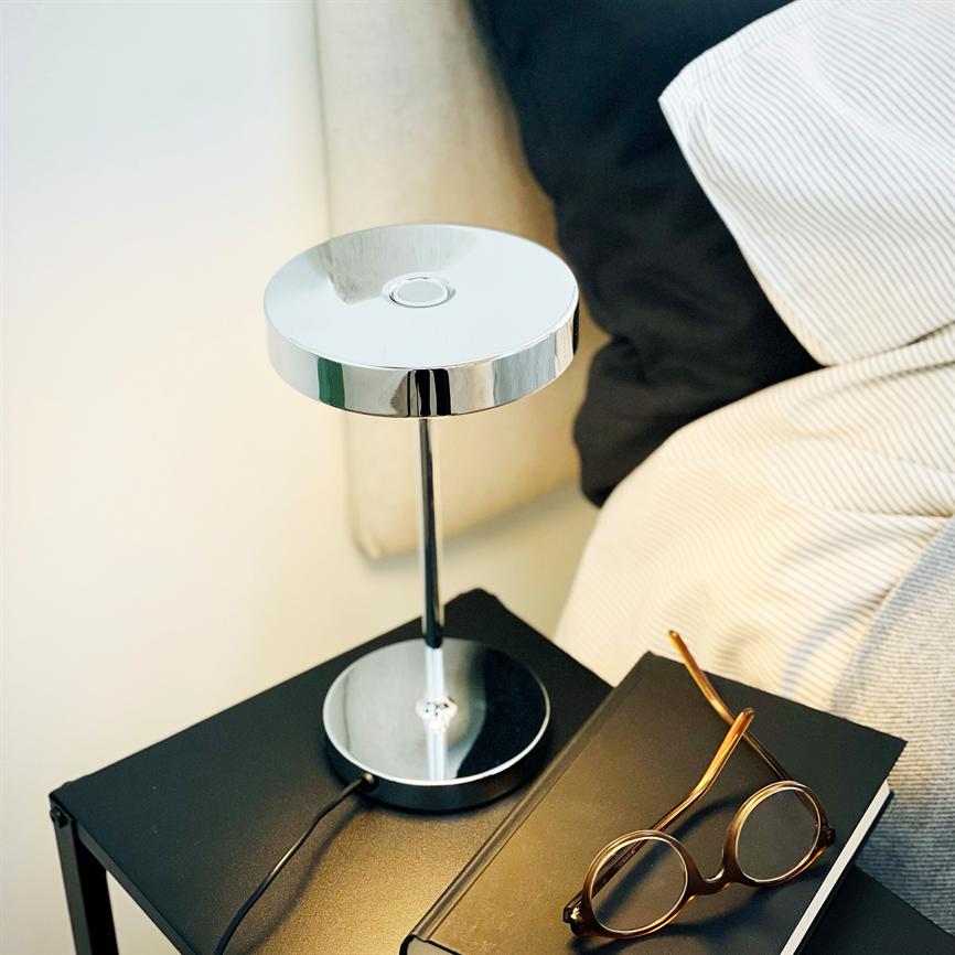 Metalbordlampe står på et sort sengebord og kaster blødt lys; en lukket notesbog og brune runde briller ligger ved siden af, foran en seng med sort pude og stribet lagen.