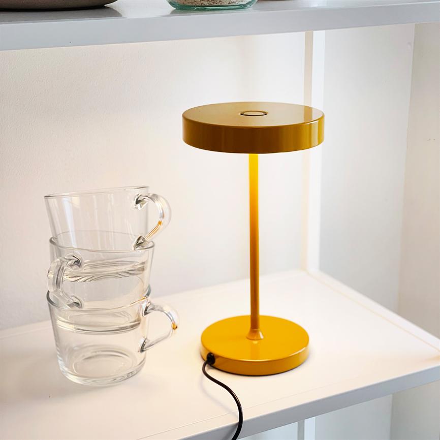 Gul bordlampe lyser blødt på en hvid hylde; ved siden af står tre stablede glaskopper, og en sort ledning løber fra lampens fod.