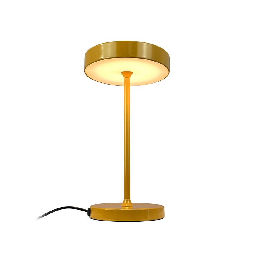 En gul bordlampe med diskformet skærm udsender varmt nedadvendt lys, stående på rund fod med slank stang og synlig sort ledning mod hvid baggrund.