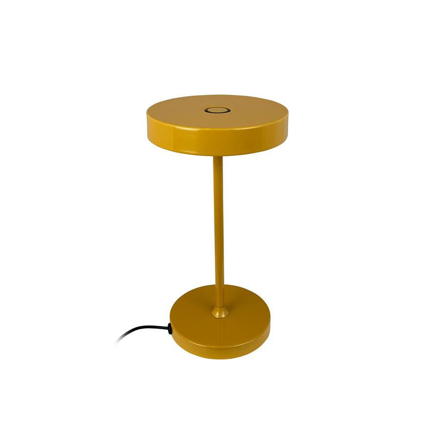 Gul metal bordlampe står isoleret på hvid baggrund; rund skærm og fod forbundet af en slank stang, sort strømkabel ligger ved basen.