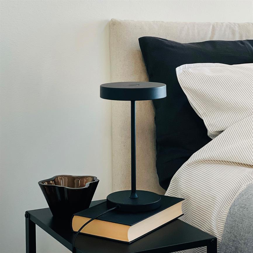 Bordlampen står placeret på en bog på et sort natbord; en brun keramikskål står ved siden af. I baggrunden ses en polstret sengegavl, sort pude og stribet sengetøj.