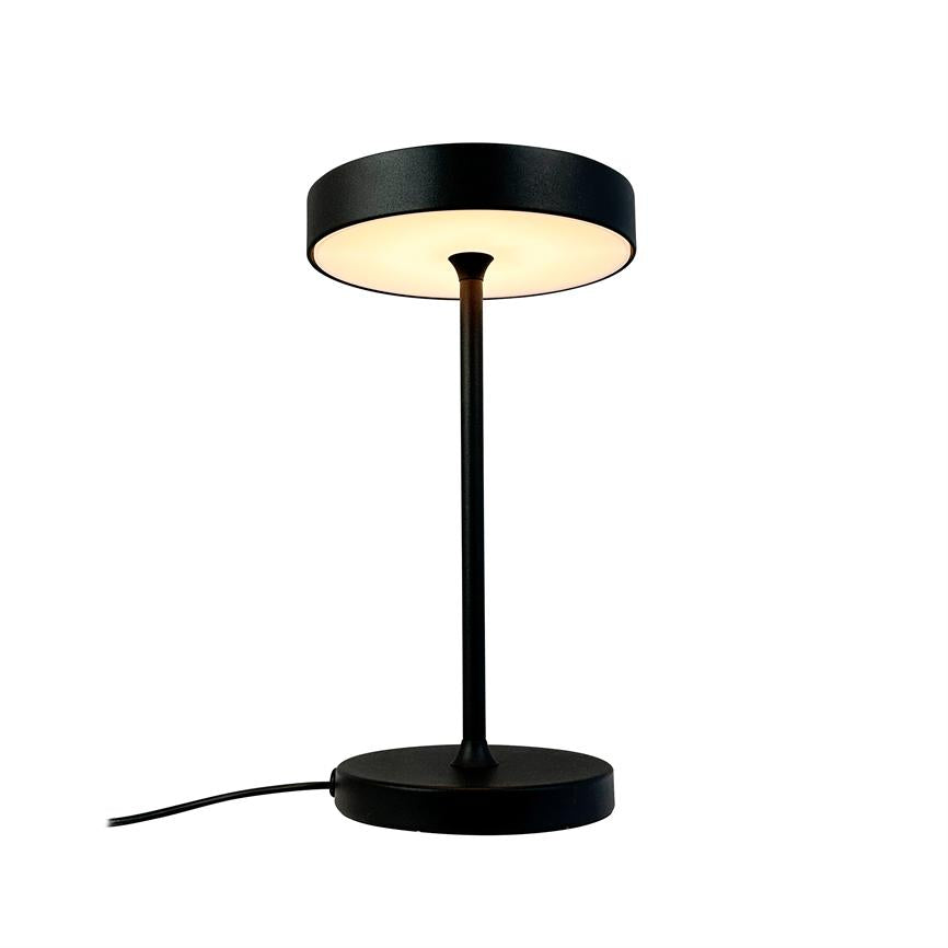 Sort, moderne bordlampe med rund skærm og slank fod lyser varmt nedad; står centreret på hvid baggrund med synligt strømkabel, minimalistisk produktfoto.