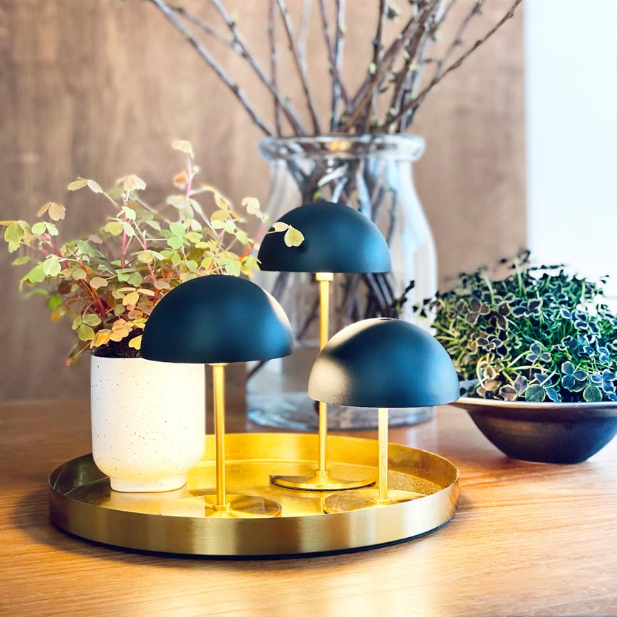 Tre små sorte kuppellamper står og kaster blødt, varmt lys på en rund guldfarvet bakke på et træbord, omgivet af potteplanter og en glasvase med kviste.