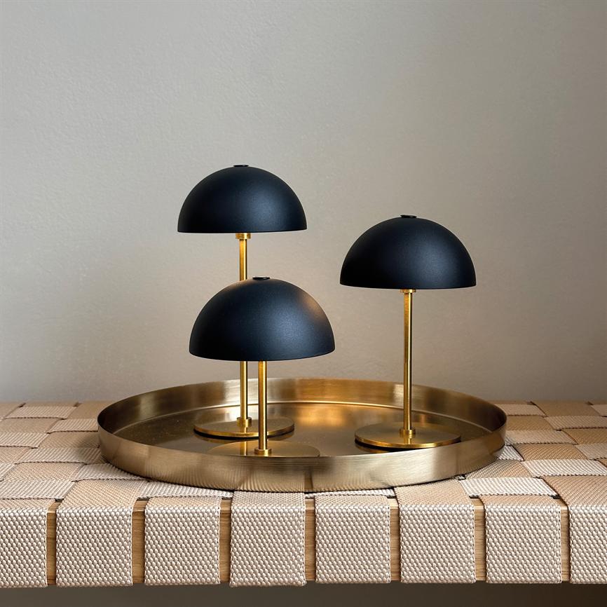 Tre små sorte kuppel-lamper står i varierende højder på en rund messingbakke, placeret på en vævet beige bænk foran en neutral, lys væg.