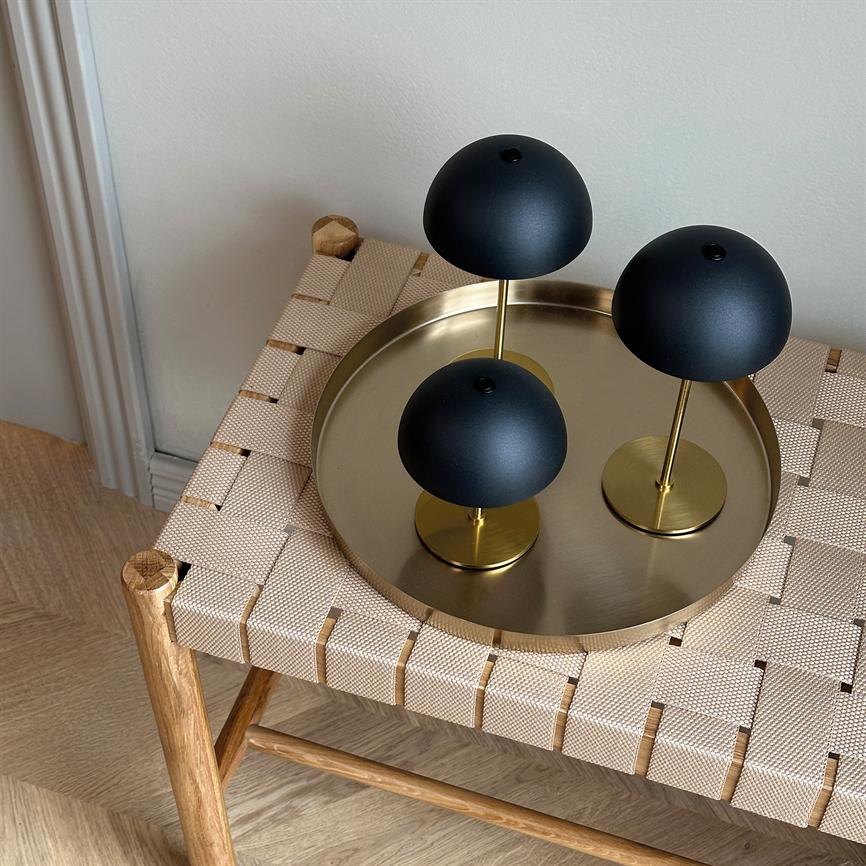 Tre sorte halvkugleformede lamper står på en rund messingbakke, som er placeret på en vævet beige bænk i et lyst hjørne med lyst trægulv og hvide vægge.