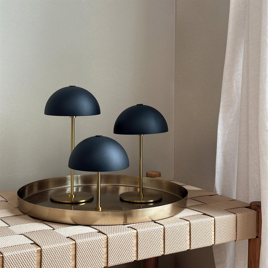 Tre matte mørkeblå skærmlamper i tre forskellige højder står på messingskaft på et rundt messingfad, placeret ovenpå en vævet bænk ved en beige væg og lyse gardiner.