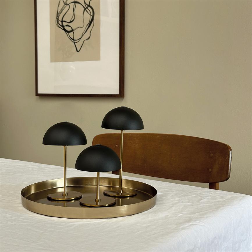 Tre sorte kuppel-lamper står på en rund messingbakke placeret på et hvidt dugdækket bord, foran en træstol og en indrammet abstrakt tegning på en beige væg.