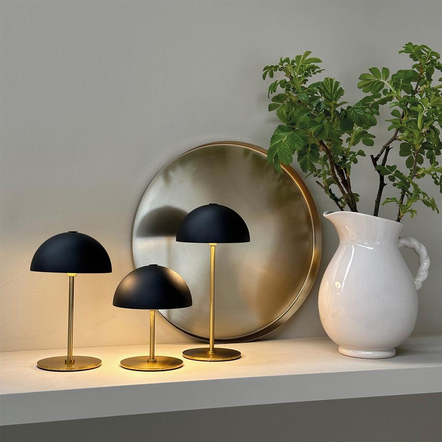 Tre sorte, guldbelagte svampeformede lamper lyser varmt på en hvid hylde; en rund messingbakke står bagved, og en hvid keramikkande med grønne grene står til højre.