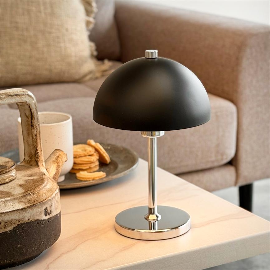 Mat sort kuppelbordslampe står på et lyst sofabord ved en keramikkanne, en kop og småkager i et lunt stuehjørne foran en beige sofa.