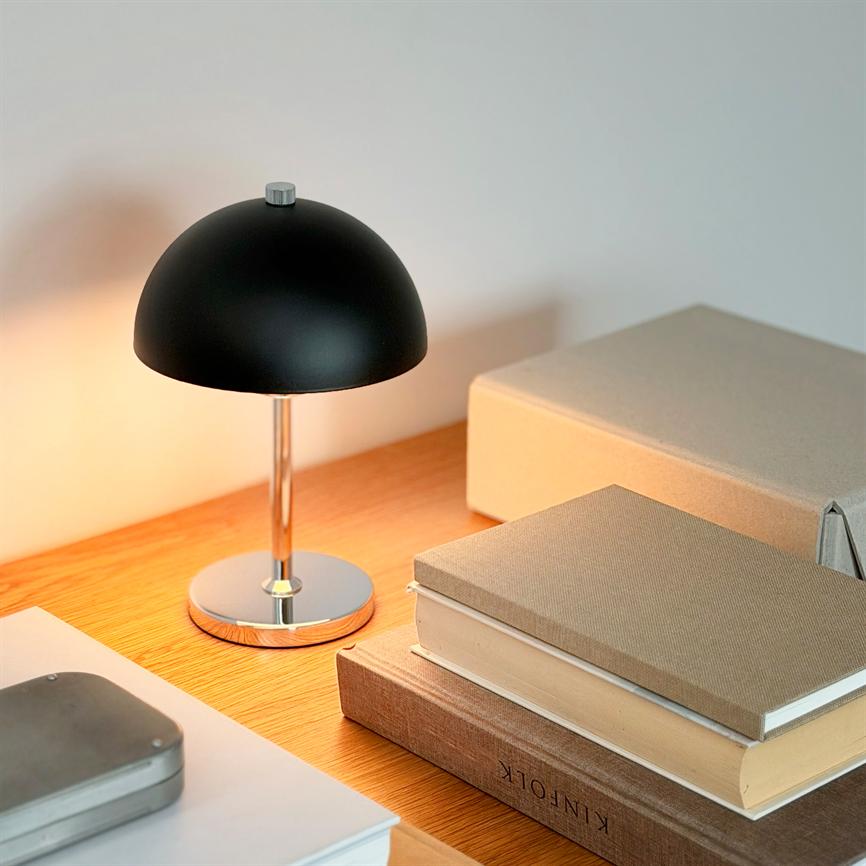 Sort, halvkugleformet bordlampe lyser blødt, kaster varme på et træbord med stabel af beige bøger og opbevaringsæsker i et roligt hjørne. Tekst i billedet: "KINFOLK".