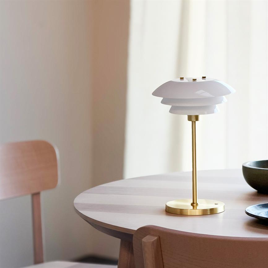 Bordlampe med hvid, lagdelt skærm og messingstang står midt på et rundt lyst træbord; omgivet af træstole, en sort skål og blødt lys gennem lyse gardiner.