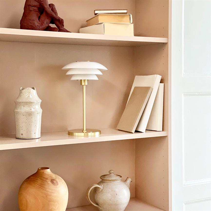 Bordlampe med guldfarvet fod og hvid skærm står midt på en beige reol; omkring den ligger stablede bøger, en lille skulptur øverst samt keramiske vaser og en tekande.