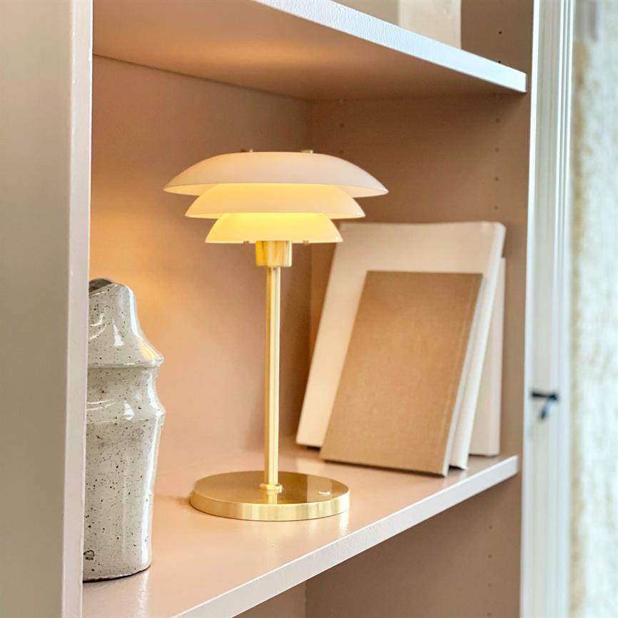 En gylden bordlampe lyser blødt; den står på en pudderfarvet hylde mellem en lysegrå keramikkrukke til venstre og to hvide/brune rammer lænet mod bagvæggen.