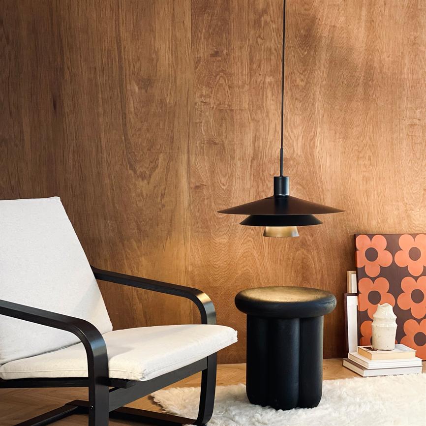 En polstret hvid lænestol med sort stel står ved siden af et rundt sort skammel under en ophængt sort pendel; træbeklædt væg, stak bøger med orange blomstermønster og hvidt pelsgulvtæppe.