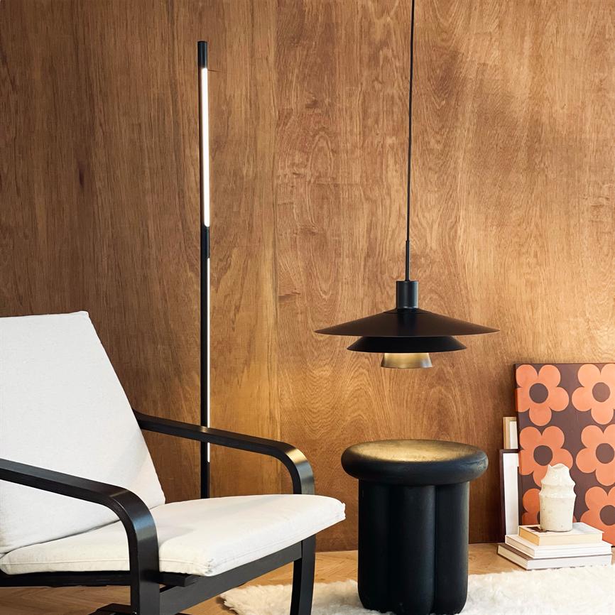 Sort pendellampe hænger over sort skammel foran en varm træbeklædt væg; en hvid polstret lænestol og en gulvlampe står ved siden af, med plakater og bøger på gulvet.