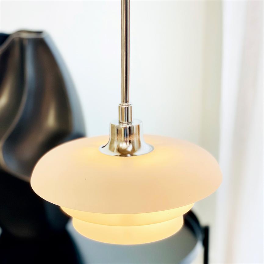 En rund, hvid ophængslampe hænger og udsender varmt lys i et lyst interiør, med et sløret sort dekorativt objekt i baggrunden og bløde hvide gardiner til højre.