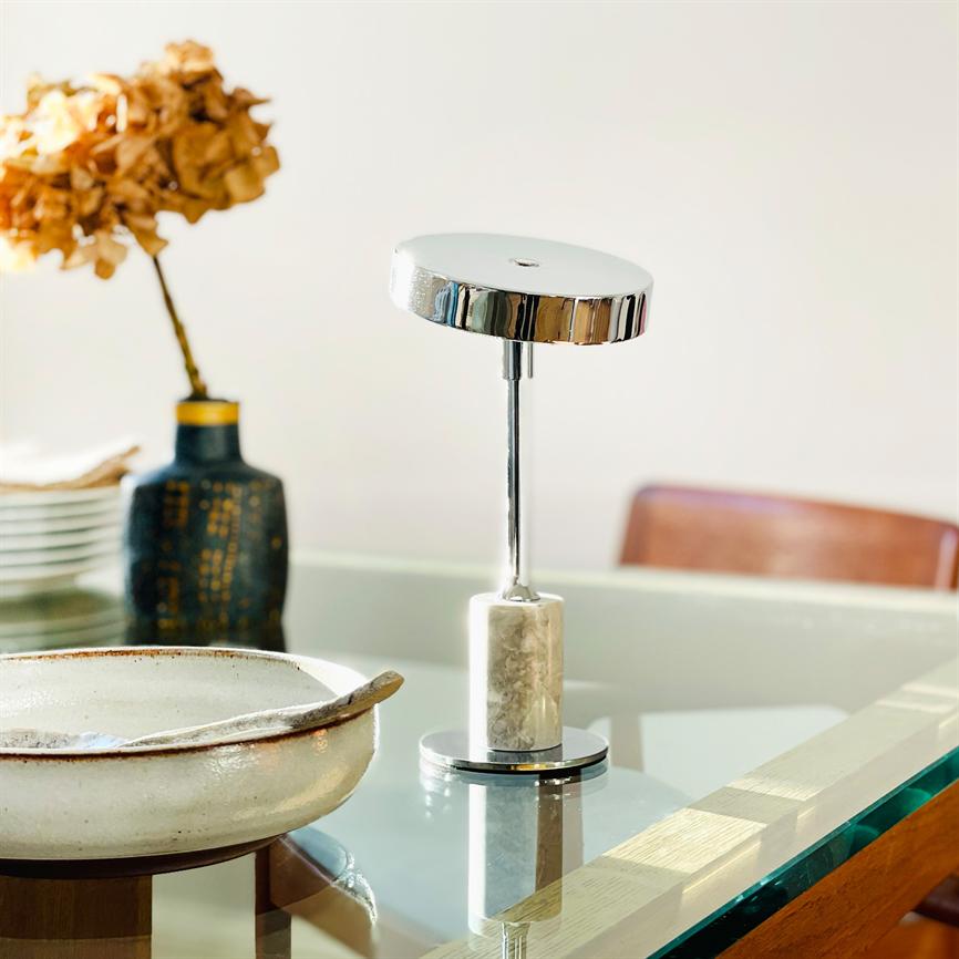 En moderne bordlampe med kromskive og marmorfod står og spejler sig på et glasbord, flankeret af en keramisk skål og en vase med tørrede blomster i spisestuen.