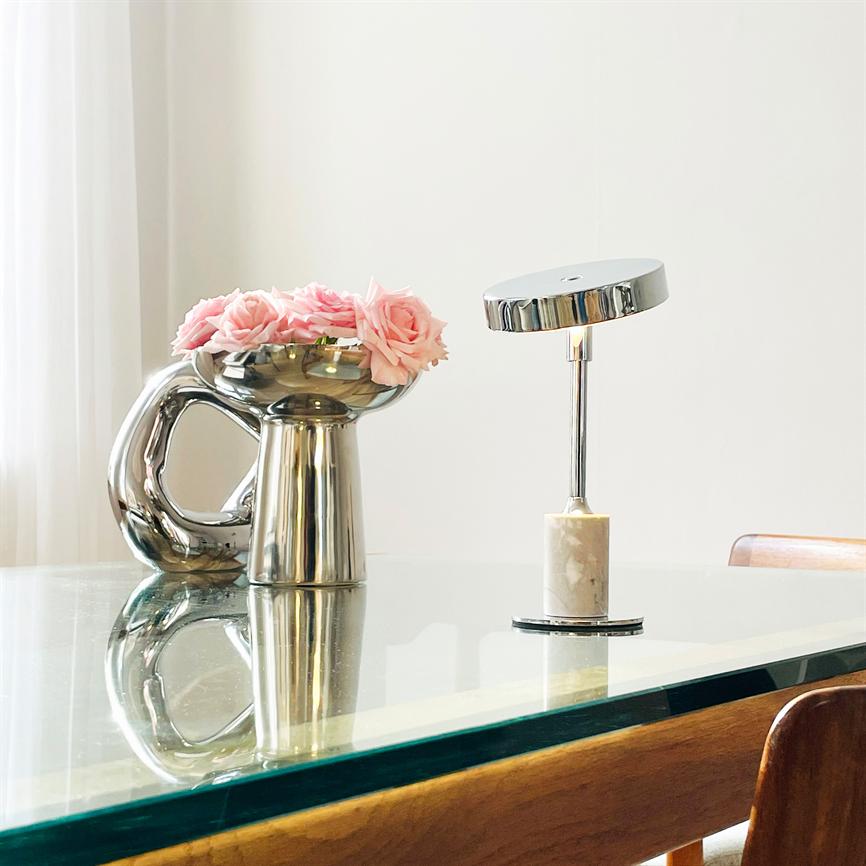 Sølvvase med lyserøde roser står på et glasbord; ved siden af en krom bordlampe med marmorfod; lyst, minimalistisk rum med hvid væg, gardin og træstolskanter.