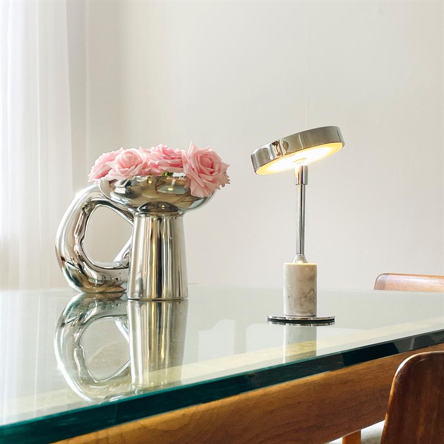 Reflekterende sølvvase med lyserøde roser står ved en tændt krom- og marmorlampe på et glasbord i et lyst, minimalistisk rum.