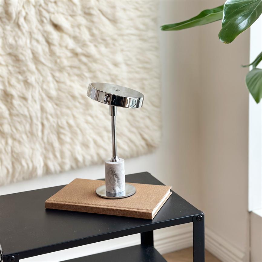 Bordlampe i krom og marmor står på en brun notesbog, hvilende på et sort metal sidebord i et lyst rum med uldagtigt vægtæppe på væggen og grønne planteblade i hjørnet.