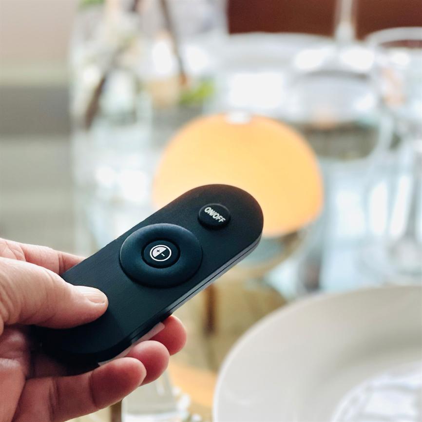 En sort fjernbetjening holdes i en hånd og peges mod en oplyst bordlampe på et dækket spisebord; der står "ON/OFF" på en af knapperne.