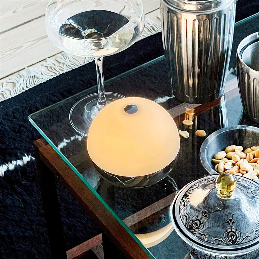 En rund, varmglødende bordlampe lyser svagt; den står på et glas sofabord omgivet af et coupeglas, metalkrus, en skål med peanuts og en mønstret lågkrukke på sort tæppe.