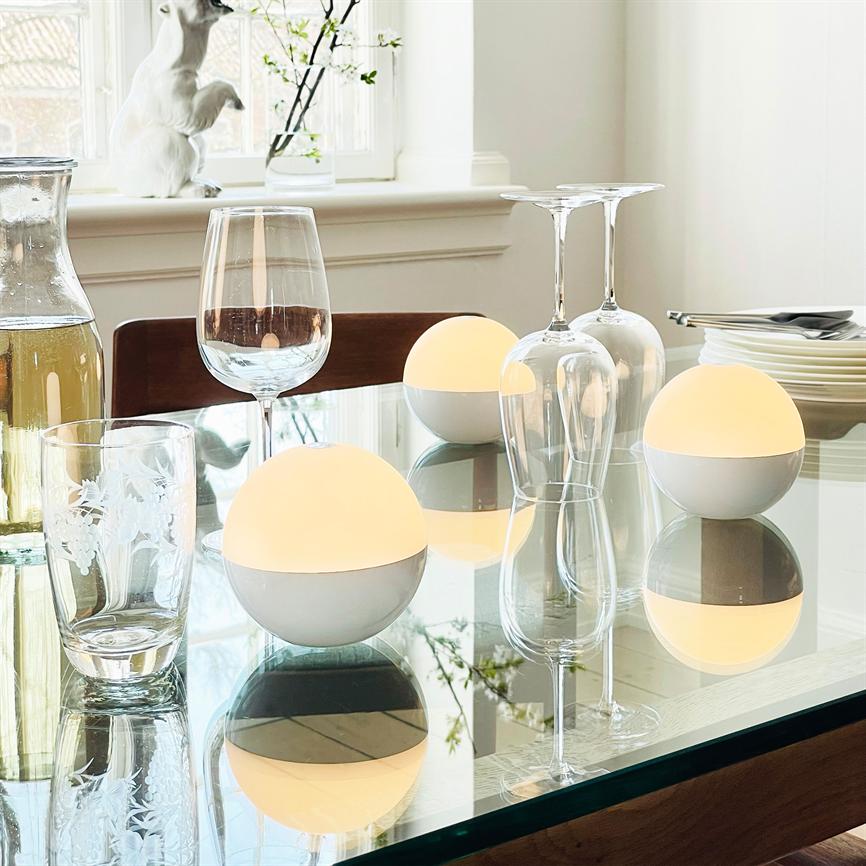 Tre kugleformede lamper udsender varm glød og spejler sig i et glasbord; omgivet af vinglas, vandkaraffel, drikkeglas og hvide tallerkener foran et lyst vindue med en lille dekorativ kanin.