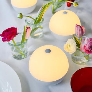 Runde, glødende bordlamper kaster blødt, varmt lys over et hvidt dækket bord, omgivet af små glasvaser med farverige tulipaner og ranunkler.