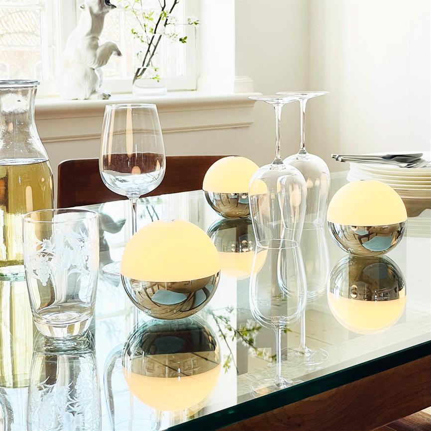 Tre halvkugleformede bordlamper med blanke metalunddele og gule overflader udstråler lys og spejler sig på et glas spisebord omgivet af vinglas, karafler, tallerkener og et vindue med en hvid kaninfigur.