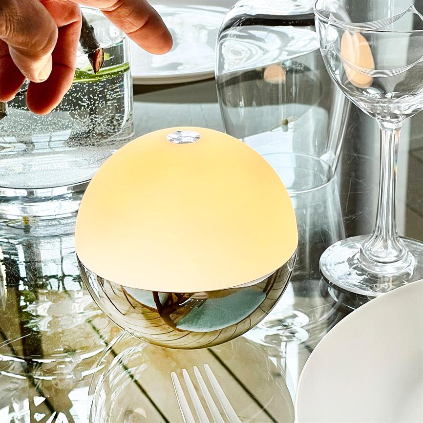 En rund bordlampe lyser varmt; den står på et spejlblankt glasbord, omgivet af vinglas, en tallerken, gafler og et glas med en hånd, der holder en plantegren.