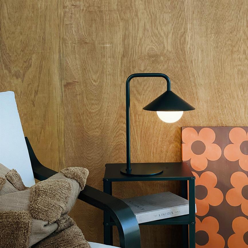 Bordlampe: sort lampe med rund skærm lyser blødt over et natbord; står foran en træbeklædt væg ved en lænestol med tæppe og et orange blomstermaleri. Tekst: "THE KINFOLK TABLE".