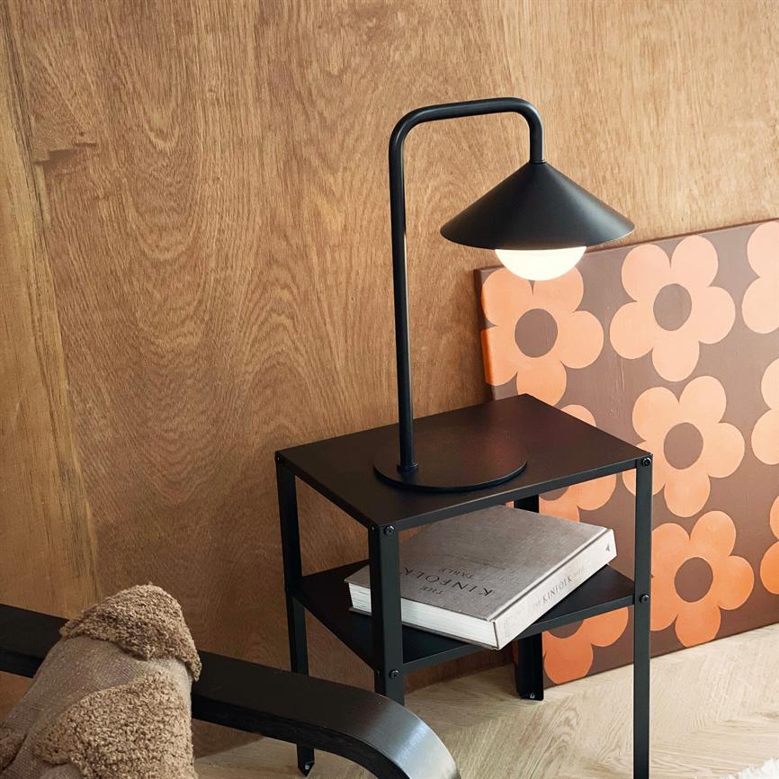 Et sort metal sidebord bærer en tændt sort bordlampe; på nederste hylde ligger bogen "KINFOLK"; omgivet af en træbeklædt væg og et orange blomsterlærred.