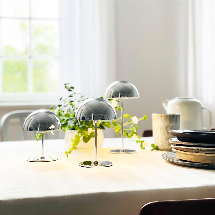 Tre små kromlamper står tændte på et hvidt dækket bord, spejler rummet; grøn potteplante, stak tallerkener og lyse gardiner ved vindue i baggrunden.