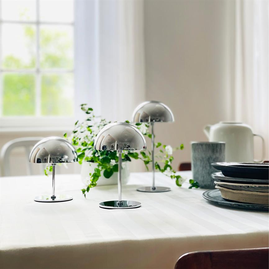 Tre små kromlamper står på et hvidt, dugdækket bord; de reflekterer rummet, omgivet af en grøn potteplante, en stak tallerkener, en kande og blødt dagslys fra et vindue.