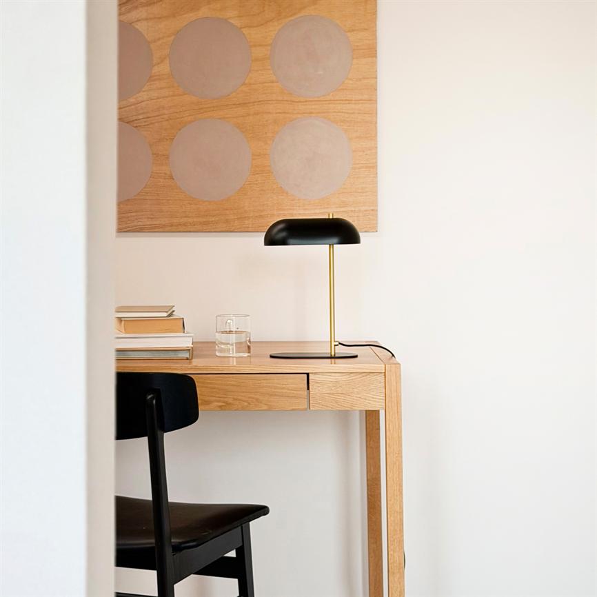 Et træskrivebord står mod hvid væg med en sort bordlampe med guldfarvet stander, bøger og et glas; et træpanel med lyse cirkler hænger bagved, en sort stol delvist synlig.