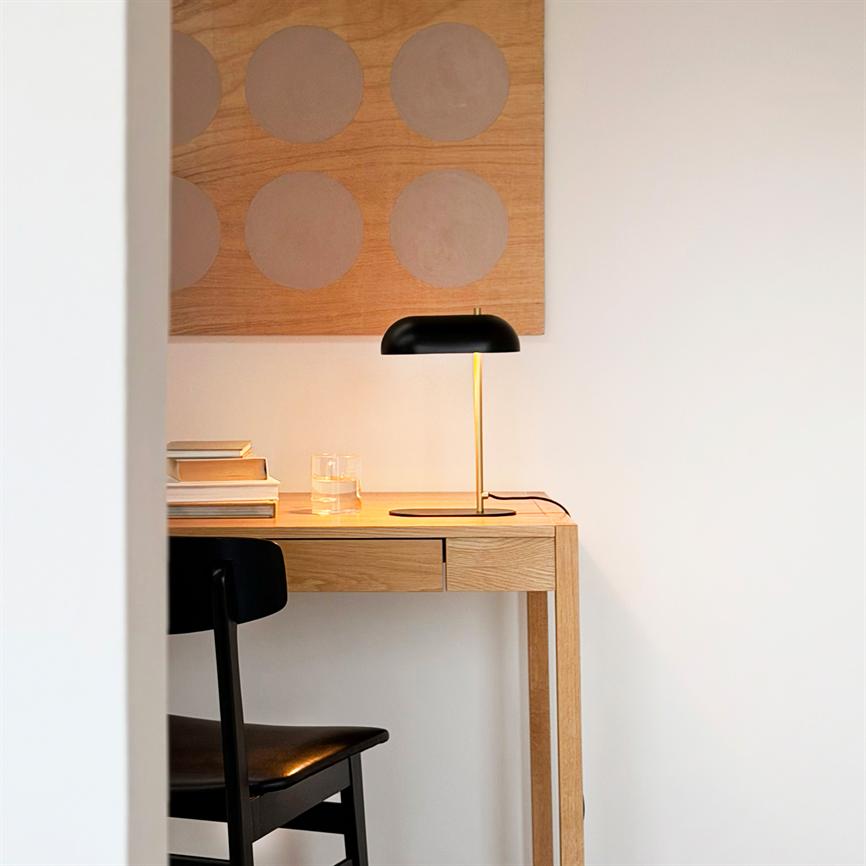 Skrivebordet er oplyst af en sort bordlampe med messingstang; på bordet ligger en bogstak og et glas. Kontekst: lyst, minimalistisk rum med sort stol og trævægkunst med cirkler.