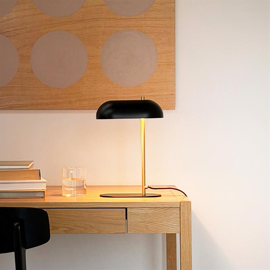 Sort og guldfarvet bordlampe oplyser et lyst egetræsskrivebord, hvor et glas vand og en stak bøger ligger; beige væg med rund mønstret træplade i baggrunden.