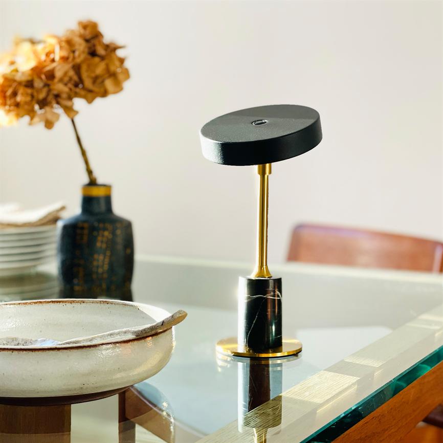 Lille moderne bordlampe med sort rund skærm og guldfarvet stang står på et glasbord; omgivet af en keramisk skål, en blå vase med tørrede blomster og en læderstol i baggrunden.