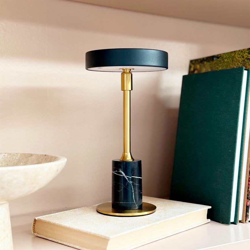 Lille designerbordlampe står på en lysebeige bog; slank messingstang med sort rundt skærm og sort marmor-fod, placeret på en hylde mellem en skål og grønne indbundne bøger.