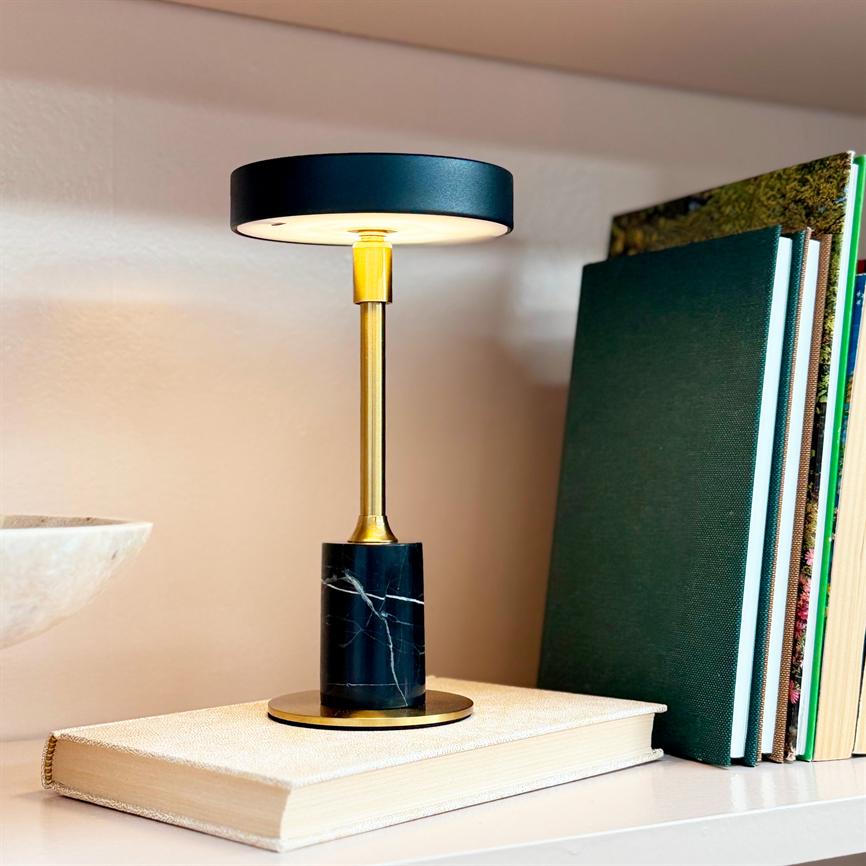 Bordlampe lyser blødt; sort rund skærm på guldfarvet stang med sort marmorfod står på en lysegrå indbundet bog på en hvid hylde ved siden af opretstående bøger.