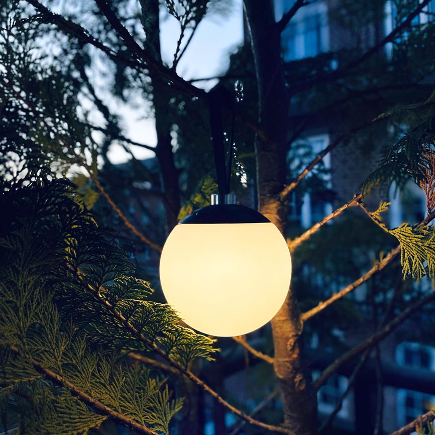 En rund, hængende lampe udsender varmt lys, hængt i en rem fra et træ; lyset oplyser nærværende nålegrene, mens sløret boligbygning og aftenhimmel ses i baggrunden.