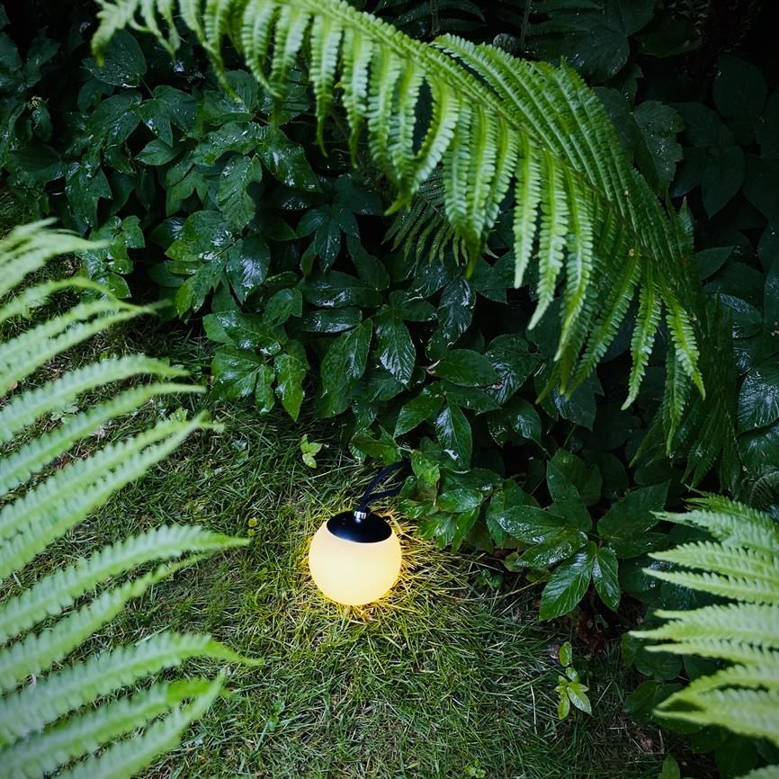 En rund solcellelampe lyser varmt på græsset, omgivet af våde grønne bregner og blanke blade i en tæt, skyggefuld havebund.