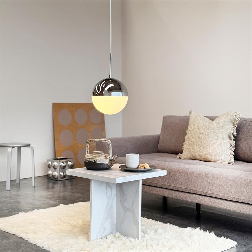 En hængende krom- og mælkehvid kuglelampe lyser over et lille marmorbord med tekande, kop og kage; omgivet af en lysegrå sofa, et blødt tæppe og minimalistisk stuemiljø.